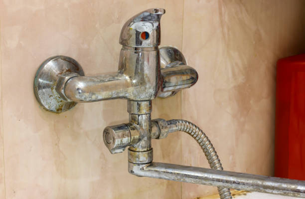 Old, corroded bathroom faucet with limescale buildup on beige tiled wall, showing neglect and poor maintenance.